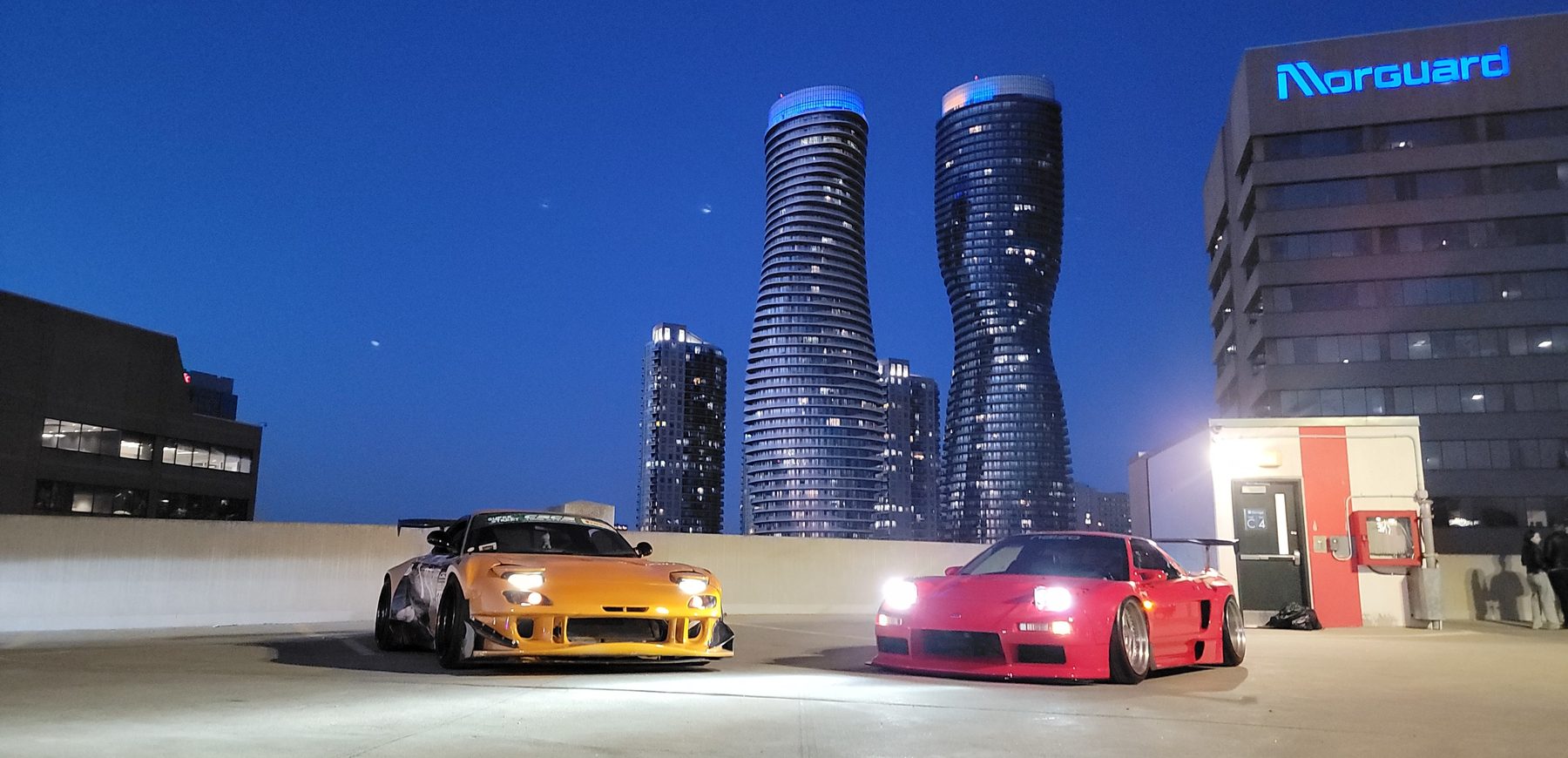 A yellow Mazda RX-7 and red Honda NSX on a rooftop with the Absolute Towers behind them at dusk