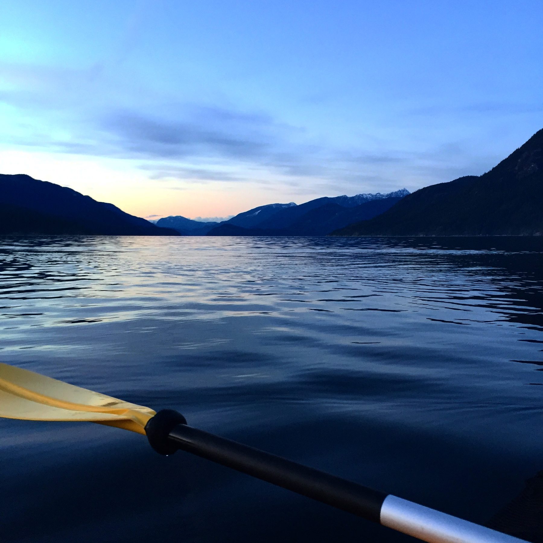 A kayak paddle skimming dark water at sunset, with mountains in the distance